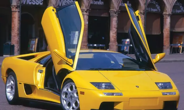Żółty Lamborghini Diablo z otwartymi drzwiami, zaparkowany na brukowanej ulicy przed budynkami z arkadami.