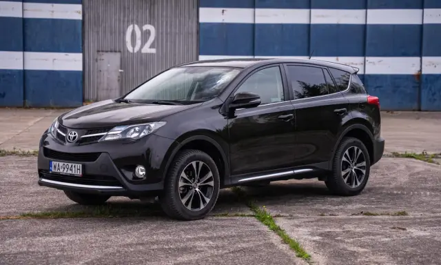Czarny, używany Toyota RAV4 z salonu, zaparkowany przed budynkiem z niebiesko-białymi pasami.