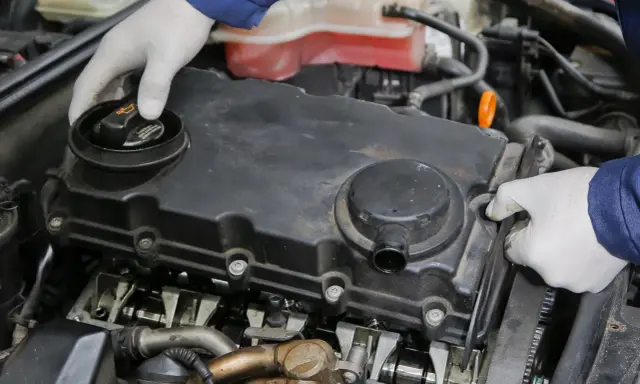 Mechanik w białych rękawiczkach sprawdza silnik, odkręcając korek wlewu oleju. Widoczne elementy silnika i układu odmy.