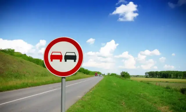 Znak zakazu wyprzedzania dla ciężarówek na tle błękitnego nieba i zielonych pól.