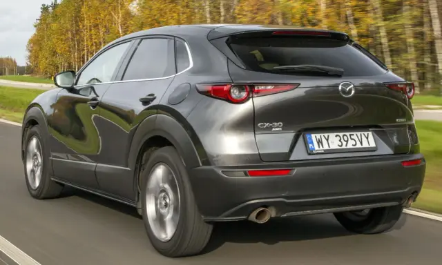 Ciemnoszara Mazda CX-30 jedzie po drodze, otoczona jesiennymi drzewami. Wady tego modelu mogą dotyczyć np. widoczności.