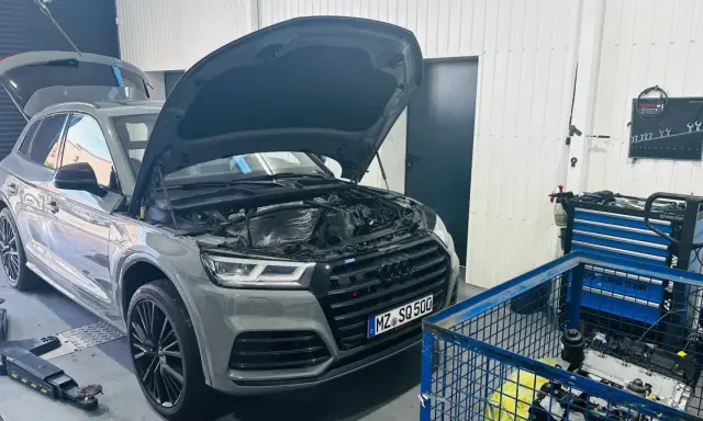 Szary Audi Q7 3.0 TDI z otwartą maską i bagażnikiem w warsztacie. Obok stoi wózek z narzędziami i silnik.