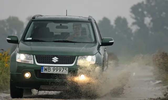Zielony Suzuki Grand Vitara 1.9 DDIS pokonuje kałużę na błotnistej drodze.