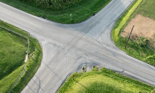 Rozjazd dróg na wsi, gdzie obowiązuje zasada prawej ręki. Wokół pola kukurydzy i zielone łąki.