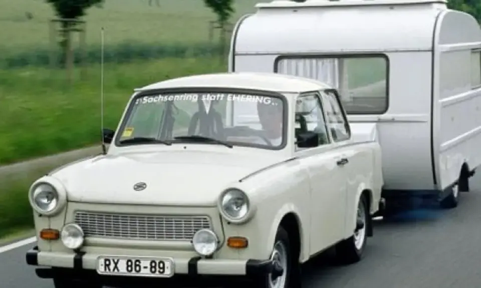 Biały Trabant ciągnie przyczepę kempingową po drodze.