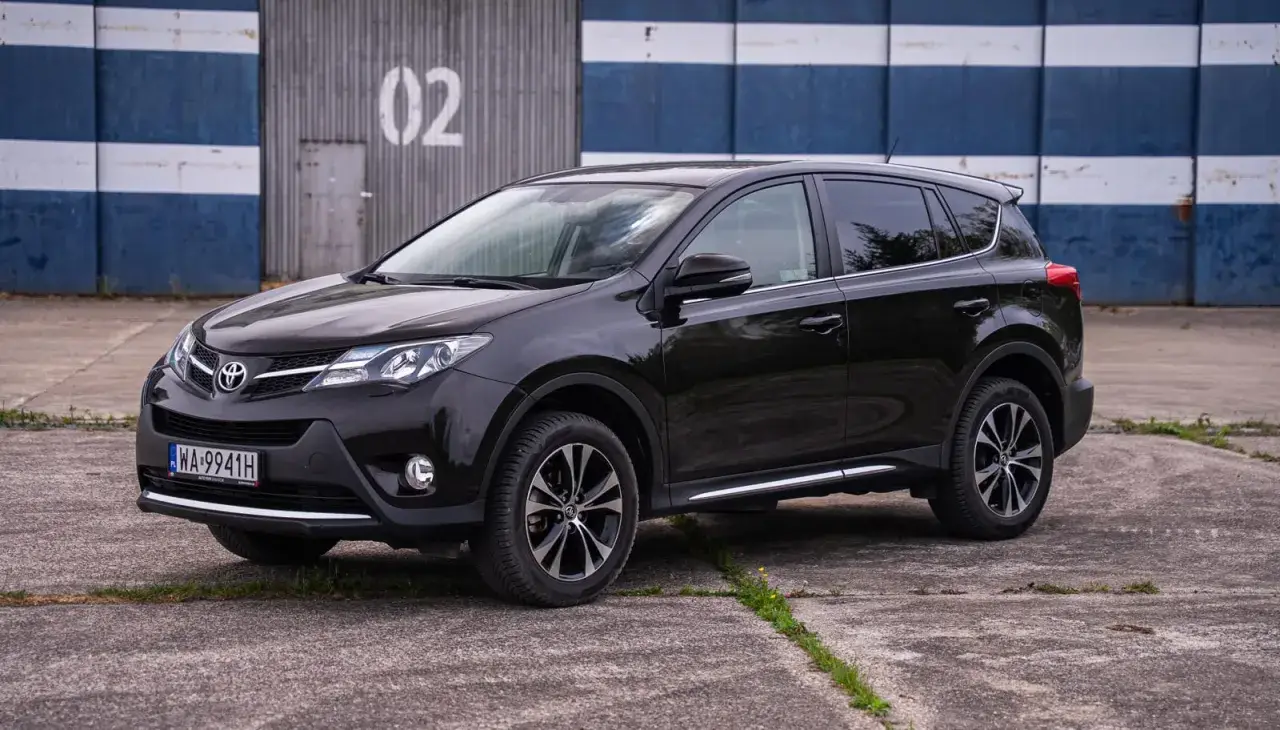 Czarny, używany Toyota RAV4 z salonu, zaparkowany przed budynkiem z niebiesko-białymi pasami.