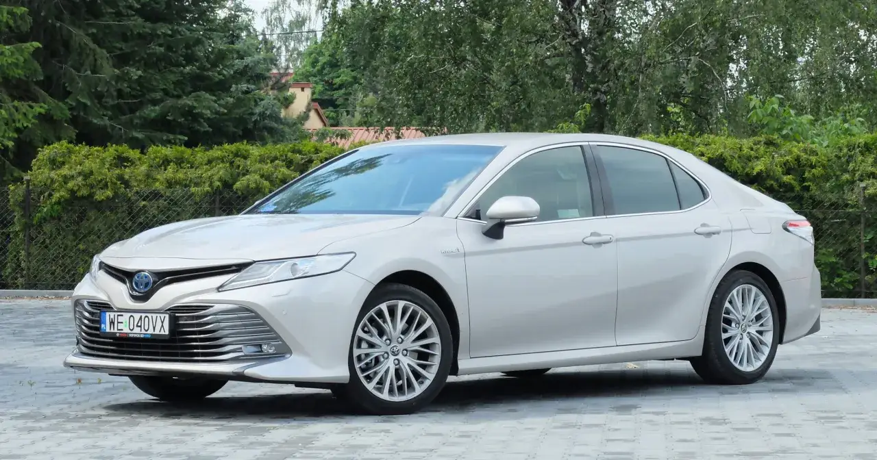 Elegancka, srebrna Toyota Camry 2024 z hybrydowym napędem prezentuje się na tle zieleni.