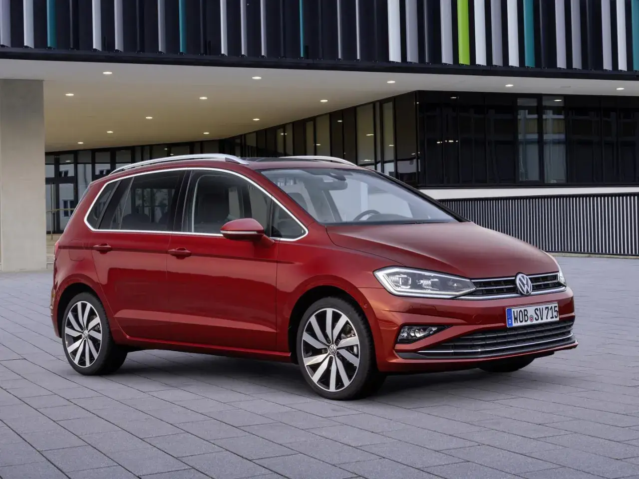Czerwony Volkswagen Golf Sportsvan przed nowoczesnym budynkiem. Wady tego modelu mogą dotyczyć np. wyciszenia kabiny.