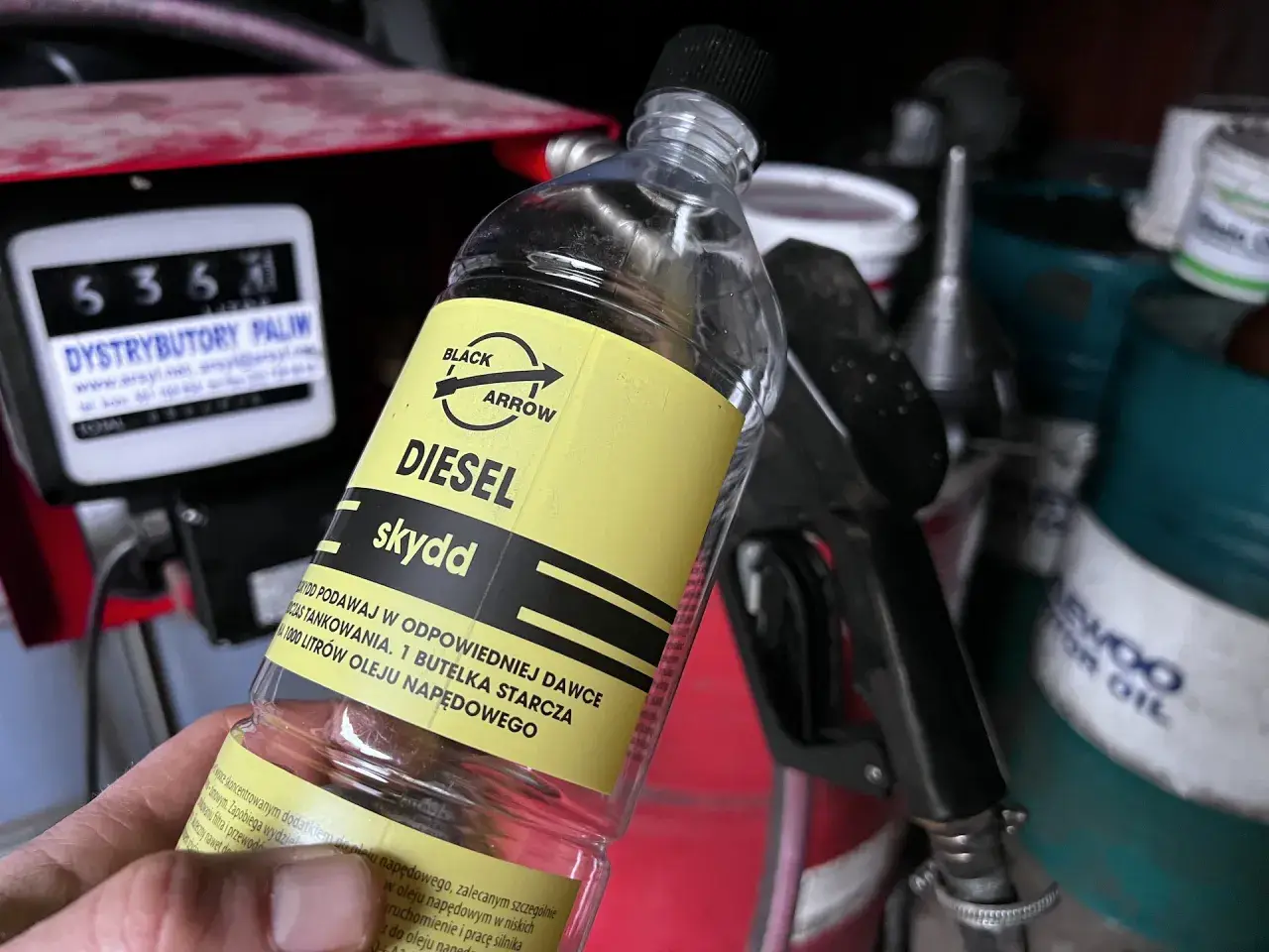 Butelka z dodatkiem do oleju napędowego, z etykietą "DIESEL skydd". W tle dystrybutor paliw i pistolet do tankowania.