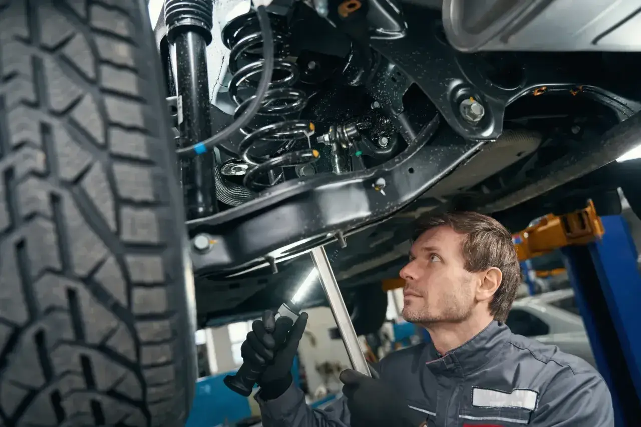 Mechanik bada podwozie samochodu, oświetlając latarką skomplikowany wielowahacz i sprężynę amortyzatora.