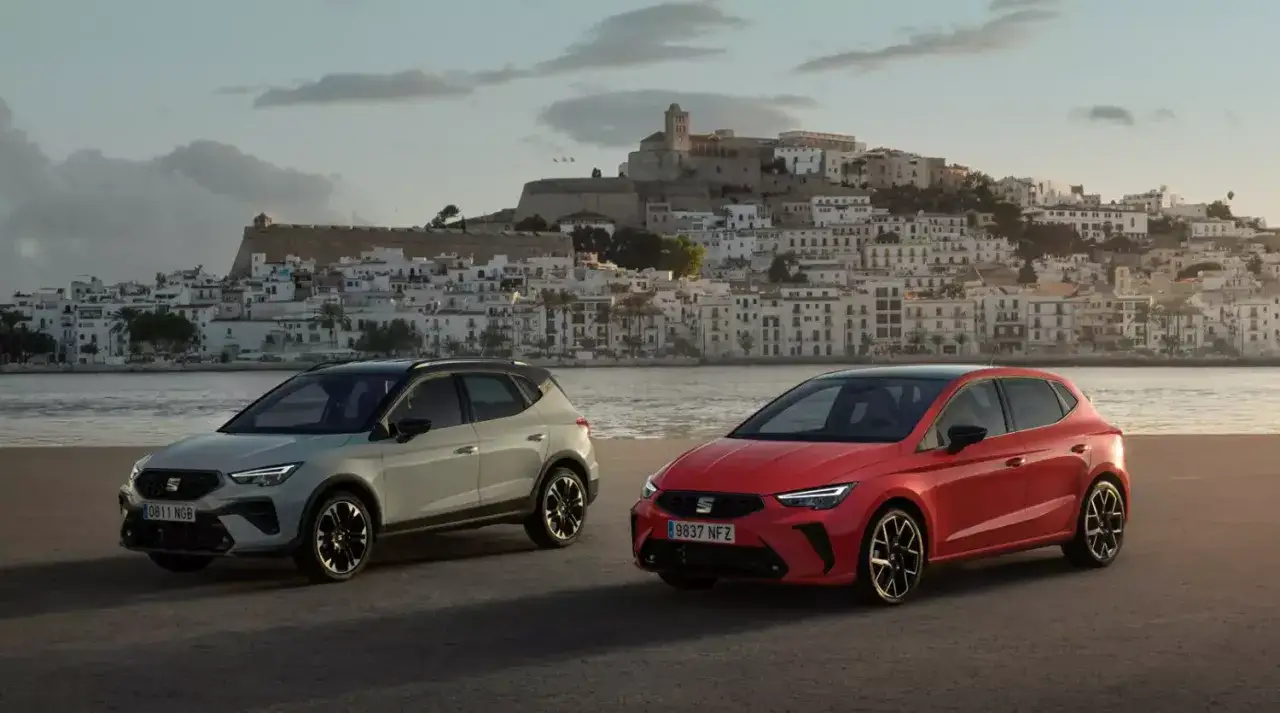 Dwa modele SEAT: czerwony hatchback i jasnoszary SUV, na tle malowniczego miasta nad morzem.