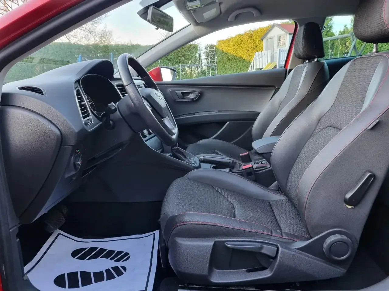 Wnętrze Seat Leon FR z czarnymi fotelami i czerwonymi przeszyciami. Widoczny kokpit, kierownica i uchwyt hamulca ręcznego.