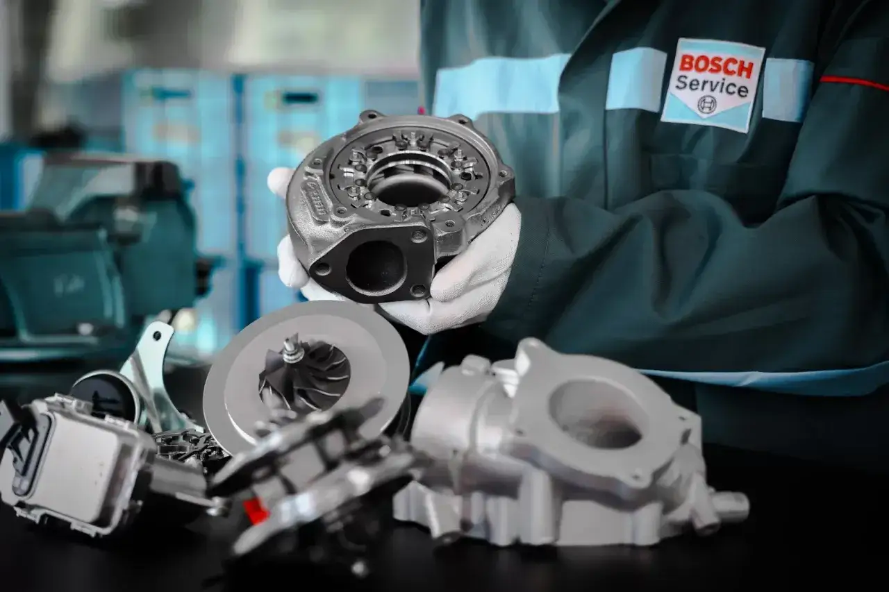 Mechanik w fartuchu Bosch Service prezentuje części turbosprężarki. Dowiedz się, ile kosztuje regeneracja turbiny.