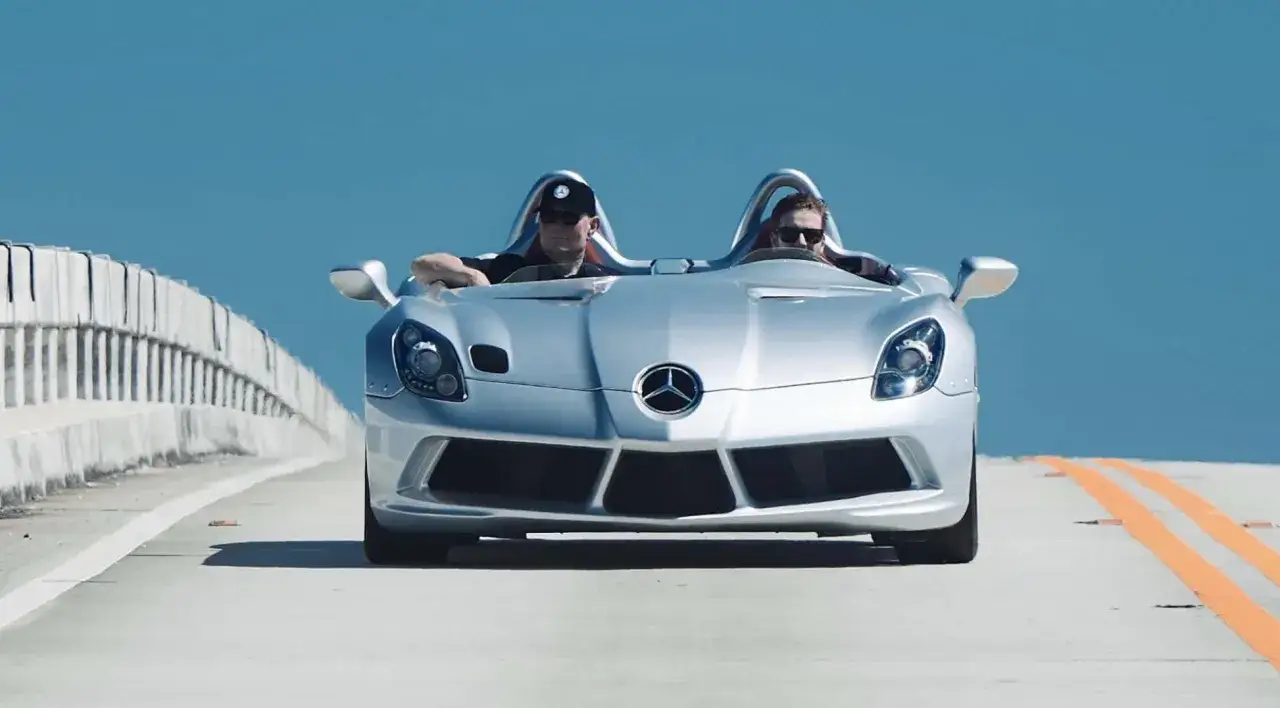 Srebrny Mercedes McLaren Stirling Moss z dwoma mężczyznami jedzie po moście, pod błękitnym niebem.