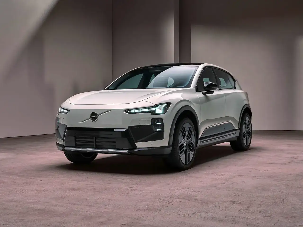Nowe Volvo EX90 w kolorze białym, z czarnym dachem i przyciemnianymi szybami, prezentuje się elegancko na tle szarej ściany.