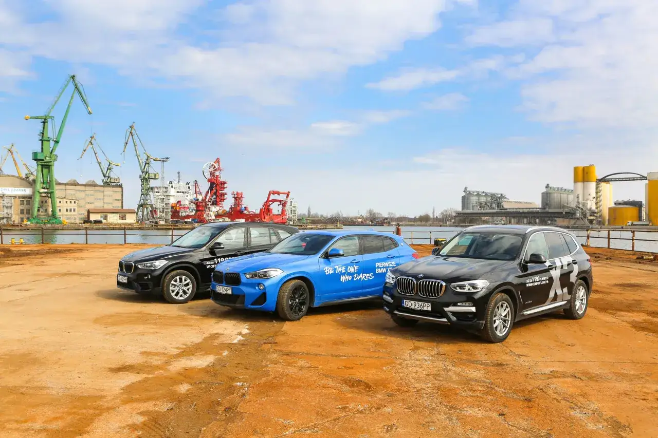 Trzy BMW X1 2025 w różnych kolorach stoją na tle portu z dźwigami.