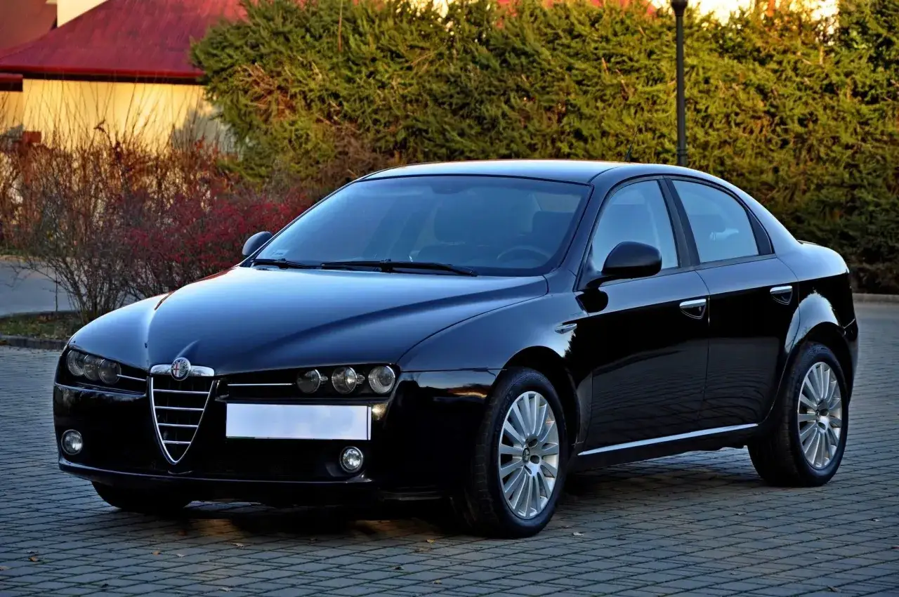 Lśniący czarny Alfa Romeo 159 1.9 JTD na brukowanej nawierzchni, z zielenią i budynkiem w tle.