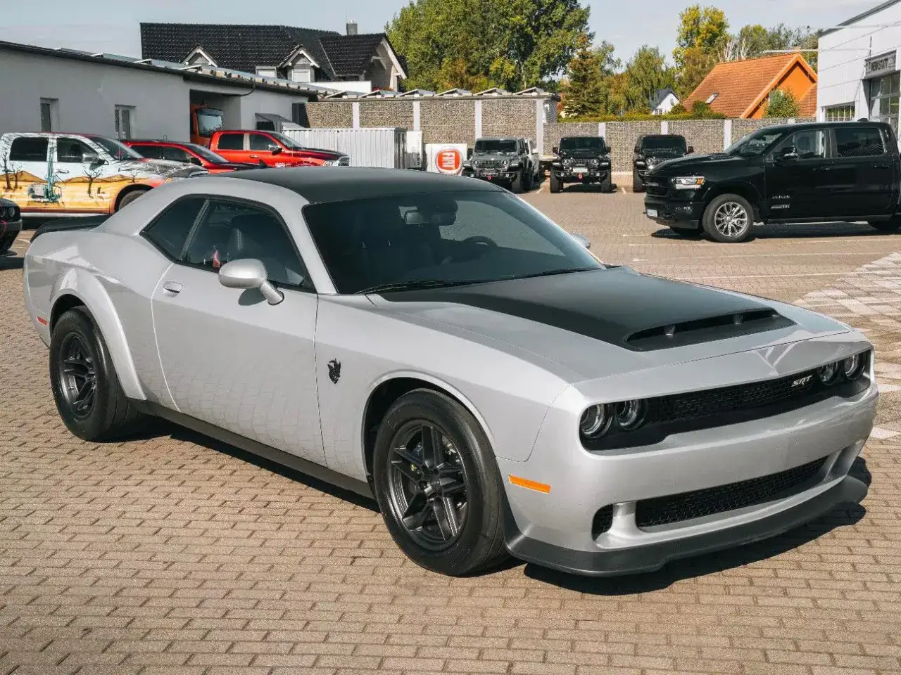 Srebrny Dodge Challenger Demon z czarną maską i dachem, gotowy do jazdy. W tle inne samochody, w tym czarny pickup.