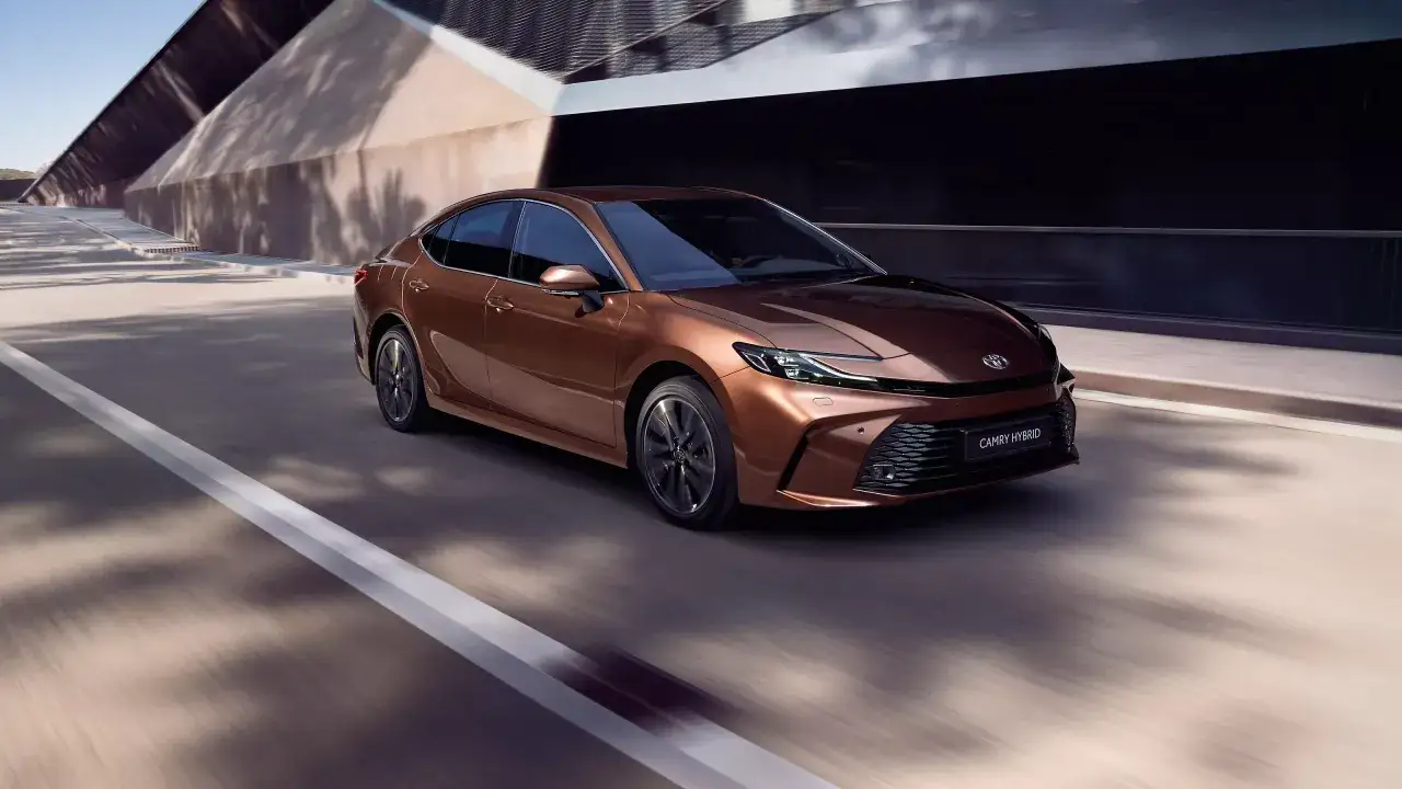 Nowa Toyota Camry 2024 w kolorze miedzianym sunie po drodze, prezentując swój nowoczesny design i hybrydowy napęd.
