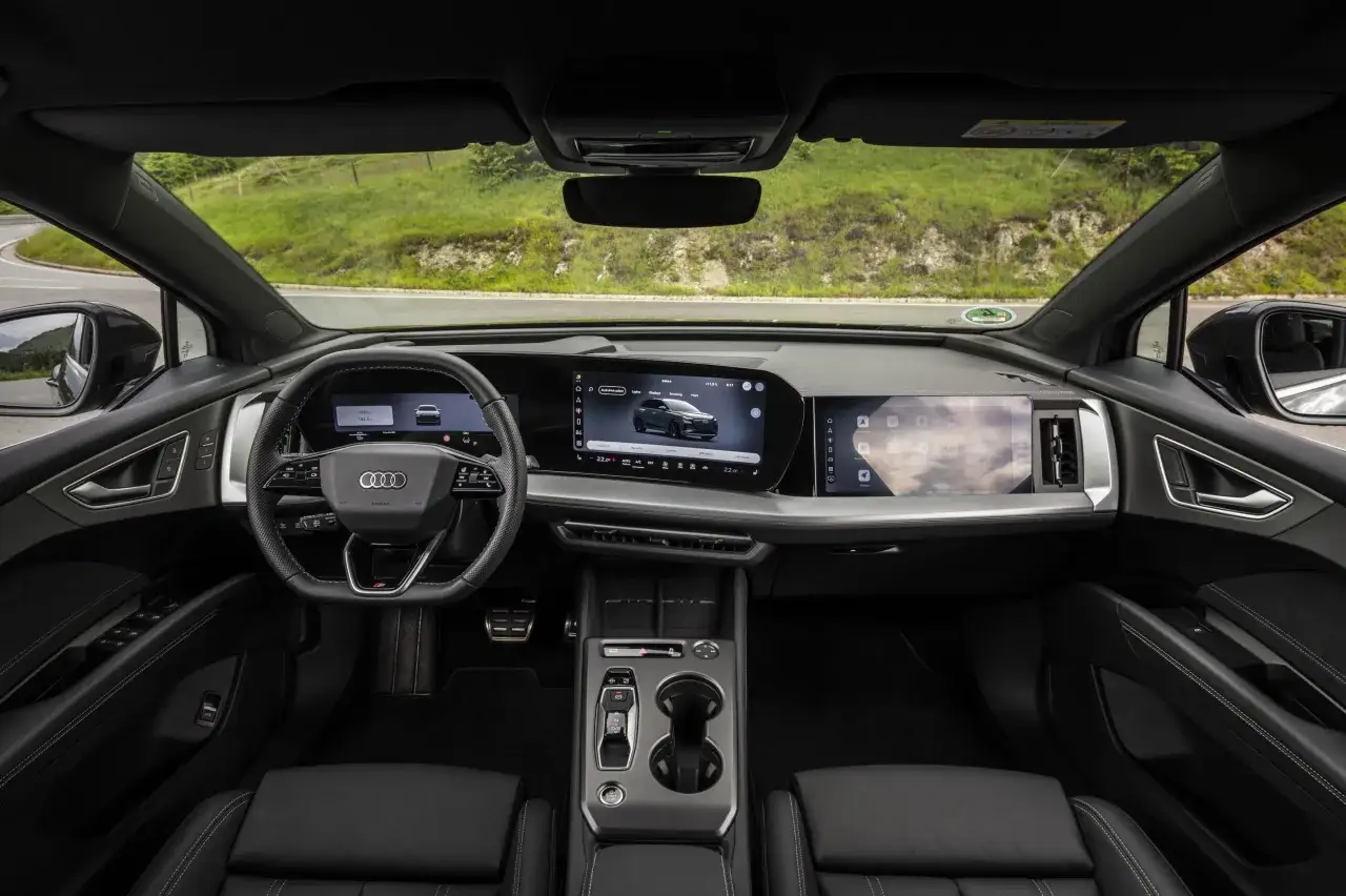 Wnętrze Audi Q4 e-tron z dwoma ekranami, kierownicą i widokiem na zielone wzgórza.
