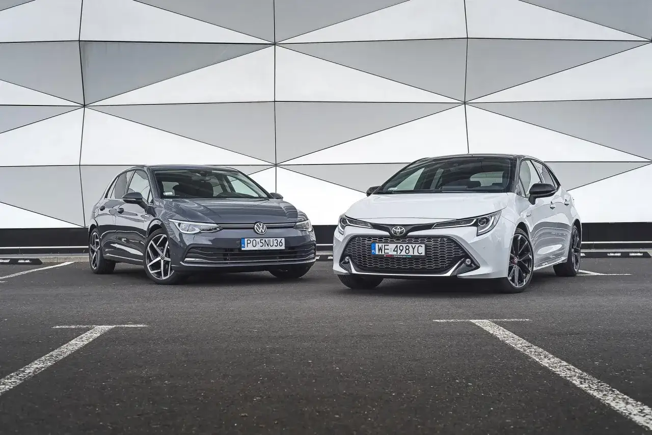 Biała Toyota Corolla GR i szary Volkswagen Golf stoją obok siebie na parkingu przed nowoczesnym budynkiem.