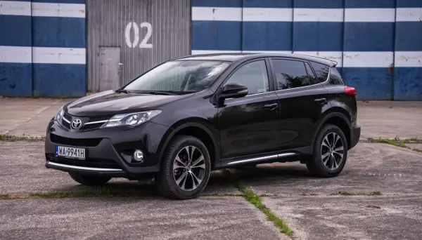 Czarny, używany Toyota RAV4 z salonu, zaparkowany przed budynkiem z niebiesko-białymi pasami.