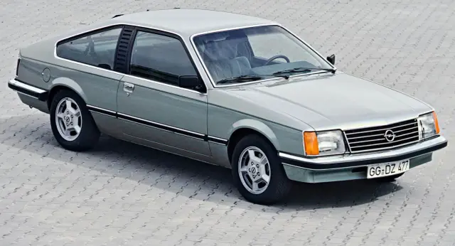 Klasyczny Opel Monza w kolorze srebrno-zielonym, z charakterystycznymi felgami i linią nadwozia, prezentuje się na tle betonowej nawierzchni.