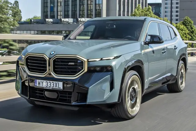 Szary BMW XM z charakterystycznym grillem i złotymi akcentami jedzie po drodze.