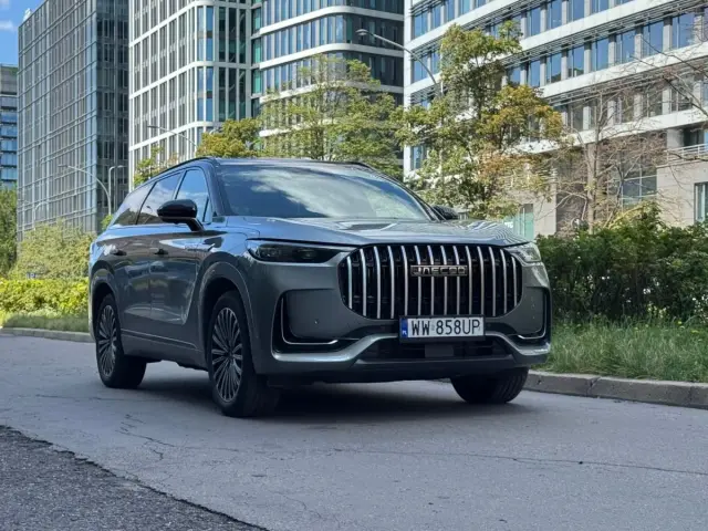Elegancki, szary SUV Jaecoo 8 z charakterystycznym grillem stoi na tle nowoczesnych budynków i zieleni.