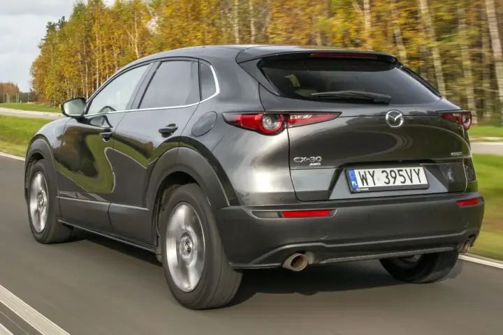 Ciemnoszara Mazda CX-30 jedzie po drodze, otoczona jesiennymi drzewami. Wady tego modelu mogą dotyczyć np. widoczności.