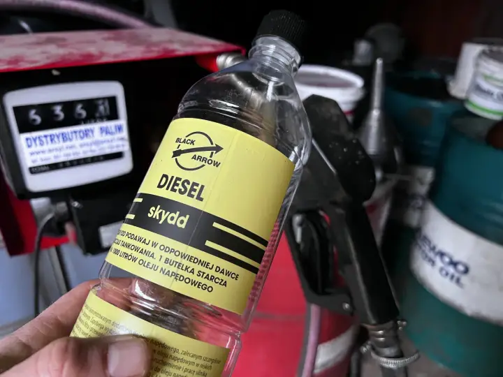 Butelka z dodatkiem do oleju napędowego, z etykietą "DIESEL skydd". W tle dystrybutor paliw i pistolet do tankowania.