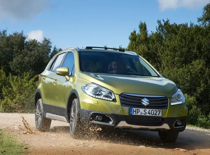 Zielony Suzuki SX4 S-Cross pokonuje teren, wzbijając kurz. Mimo że auto radzi sobie na bezdrożach, warto rozważyć jego wady przed zakupem.